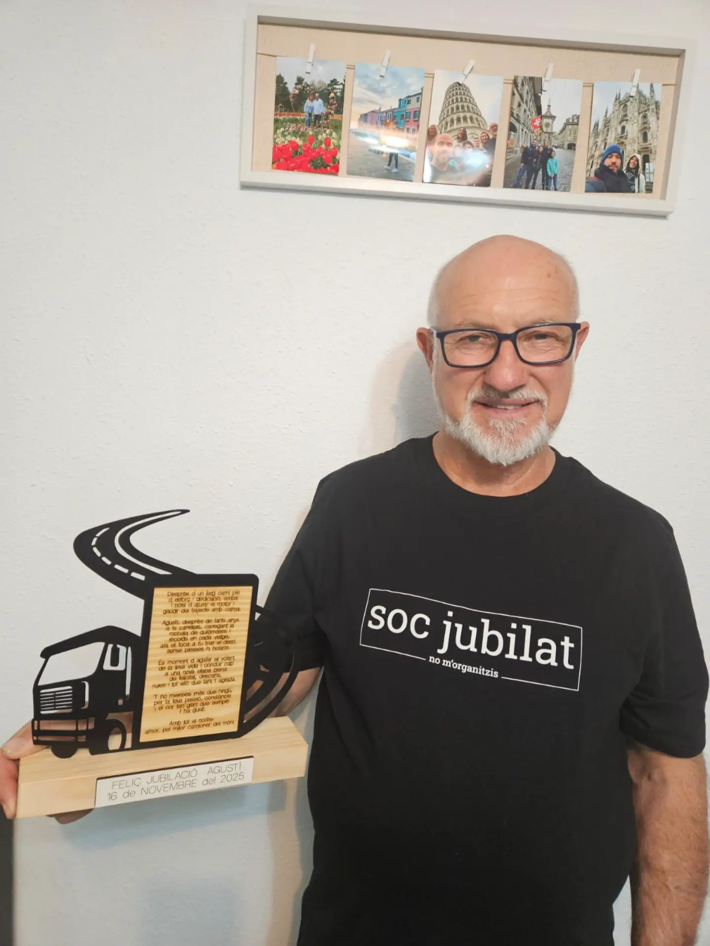 Un sonriente hombre mayor con gafas sostiene un trofeo en forma de camión, con una camiseta "soc jubilat", celebrando sus años en el transporte por carretera. Fotos en la pared de detrás.