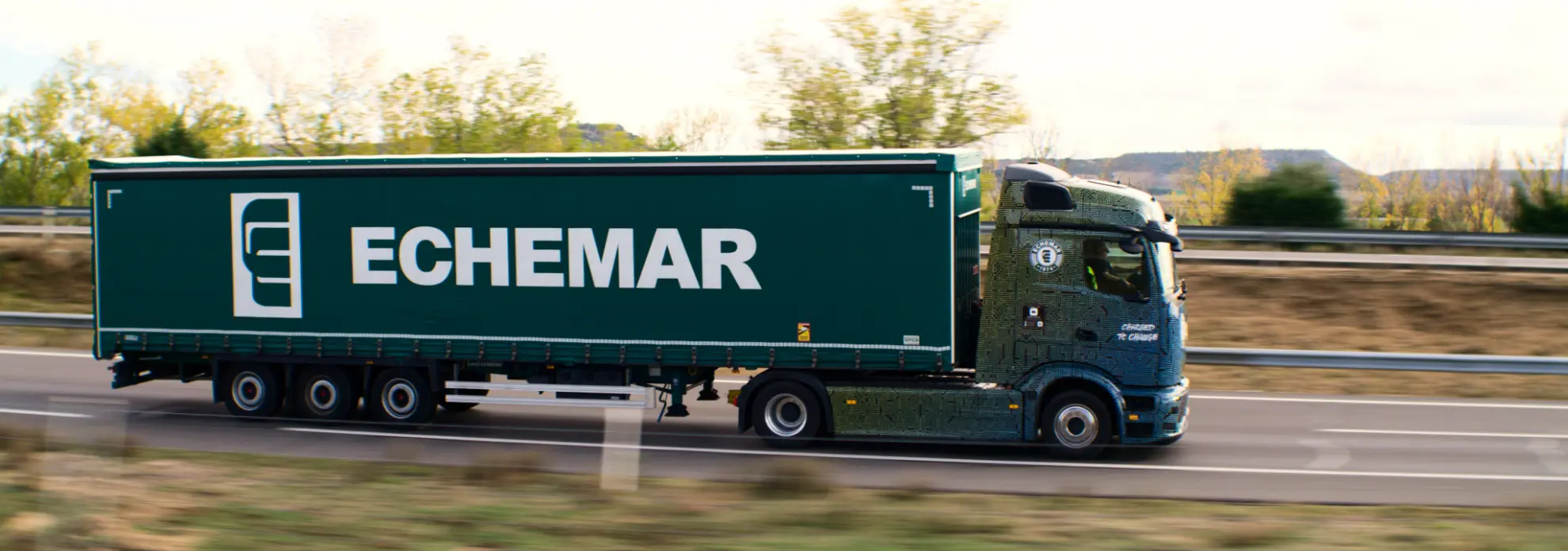 Un camión verde Mercedes-Benz Echemar, posiblemente un eActros 600, con un remolque avanza por la autopista, navegando sin problemas a través de un frondoso corredor de árboles.