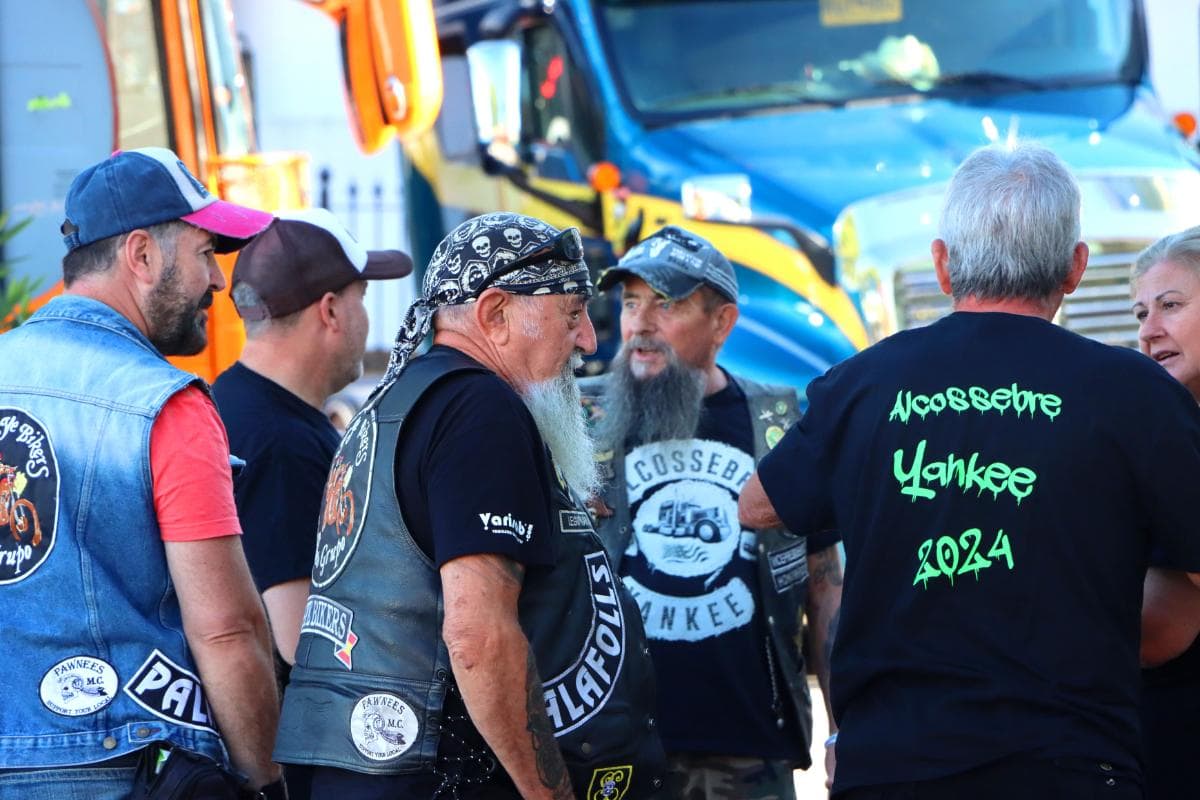 Un grupo de motociclistas vestidos de negro y de mezclilla conversan cerca de un camión azul; sobresale un hombre con una larga barba blanca. Esta descripción captura la escena vívidamente, ideal para palabras clave de SEO y necesidades de borrador automático.