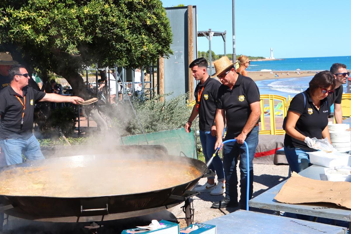 Un grupo de personas cocinando paella al aire libre en una playa en un día soleado, disfrutando del vívido paisaje y la camadería.