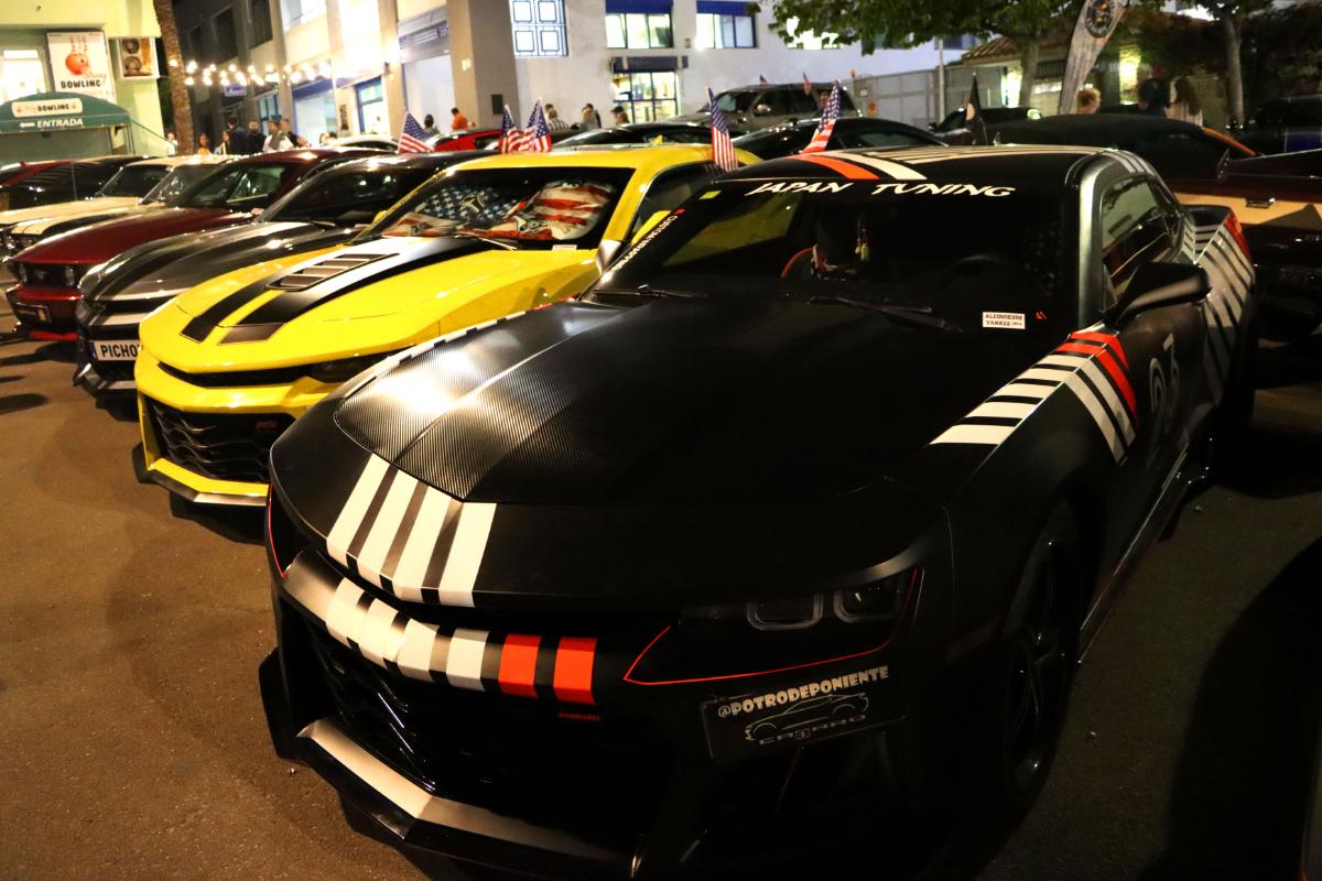 Una fila de autos deportivos, incluido un auto negro con rayas blancas y rojas, se presenta en un evento nocturno de borrador automático.