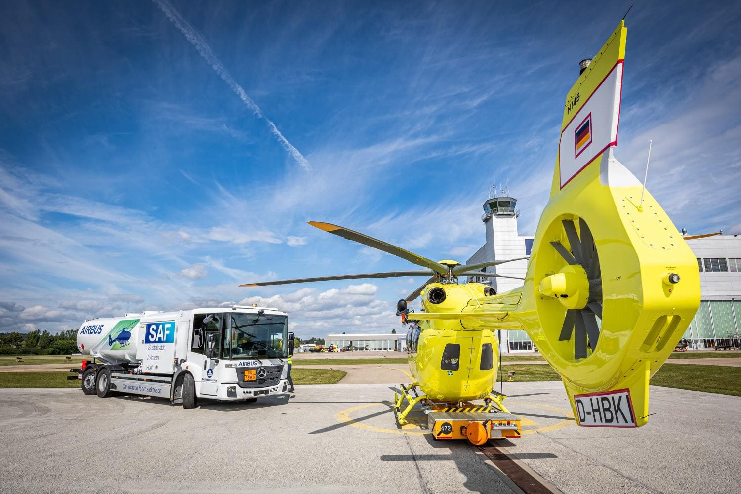 Un helicóptero amarillo y un camión de combustible se encuentran en un aeródromo con una torre de control al fondo bajo un cielo azul, asemejando la precisión de un borrador automático en movimiento.