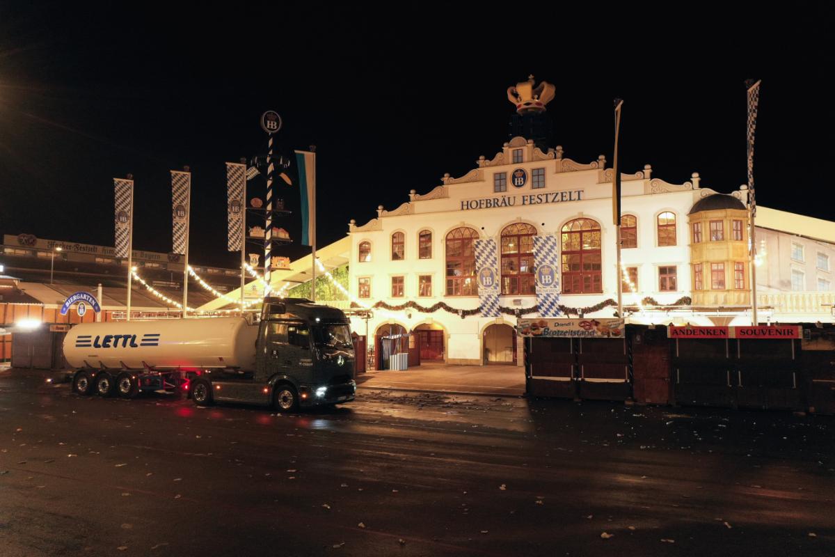 La Hofbräu Festzelt iluminada por la noche proyecta un resplandor sobre el camión de combustible estacionado frente a ella, asemejándose a una escena capturada por un borrador automático.