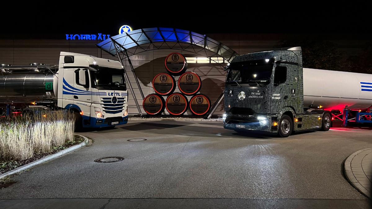 Dos camiones están estacionados frente a una cervecería por la noche, donde barriles iluminados, dispuestos con precisión, forman una llamativa forma piramidal.