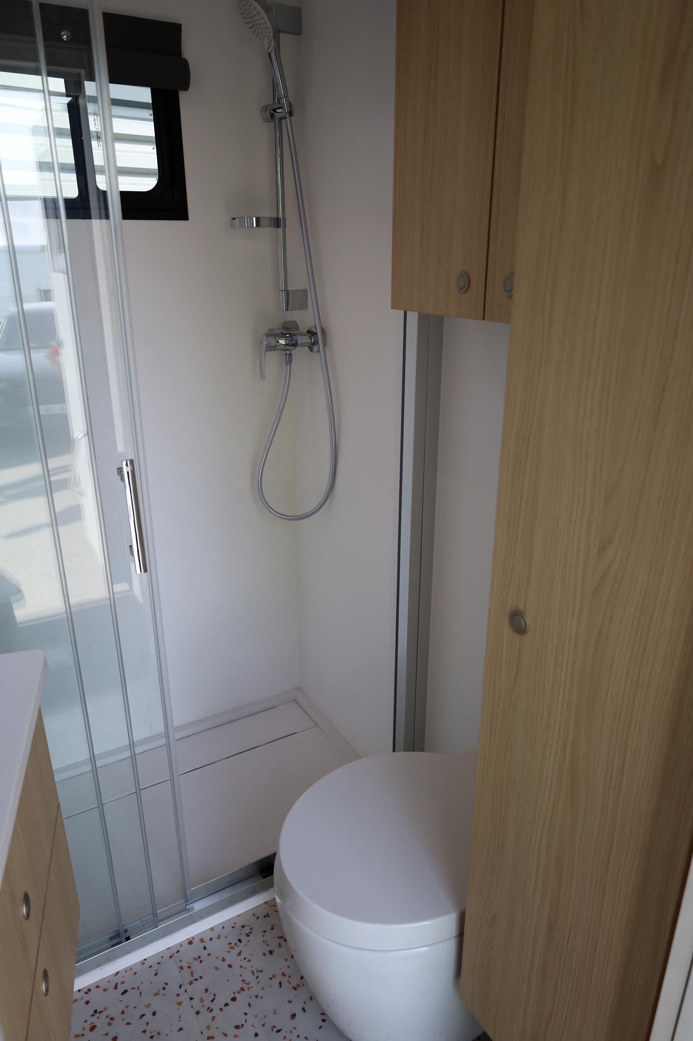 El baño compacto cuenta con ducha, inodoro y muebles de madera, con una pequeña ventana sobre la zona de la ducha para que entre luz natural. Es tan eficiente como un "borrador automático" en su diseño elegante, optimizando el espacio a la vez que mejora la funcionalidad.