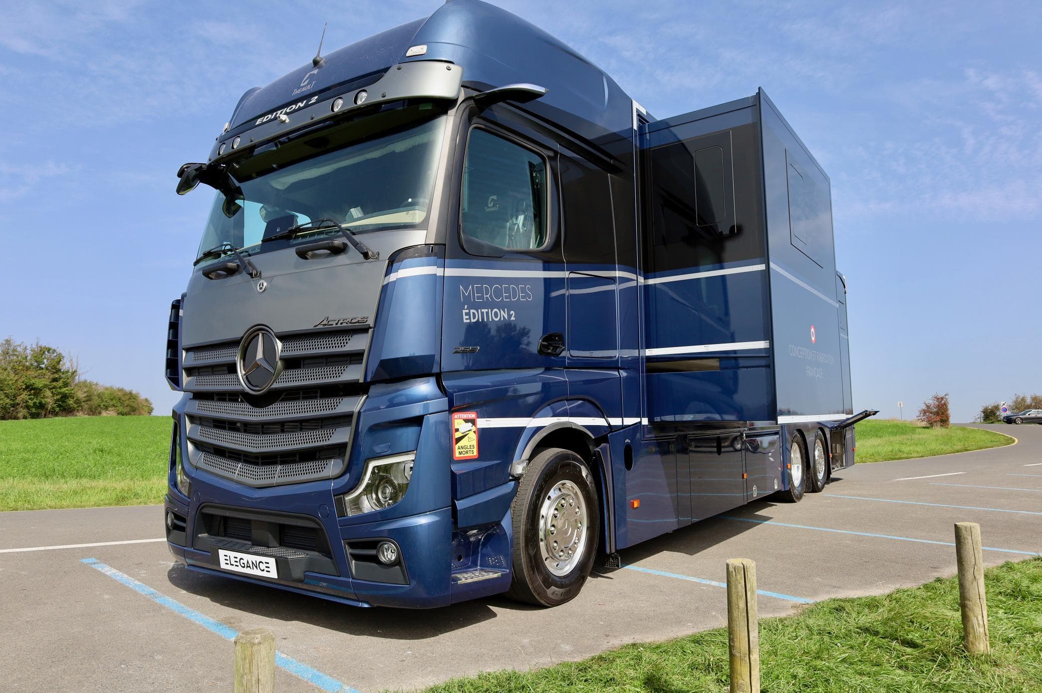 Camión Mercedes azul estacionado en una carretera al lado de un campo de hierba y un fondo de cielo azul.