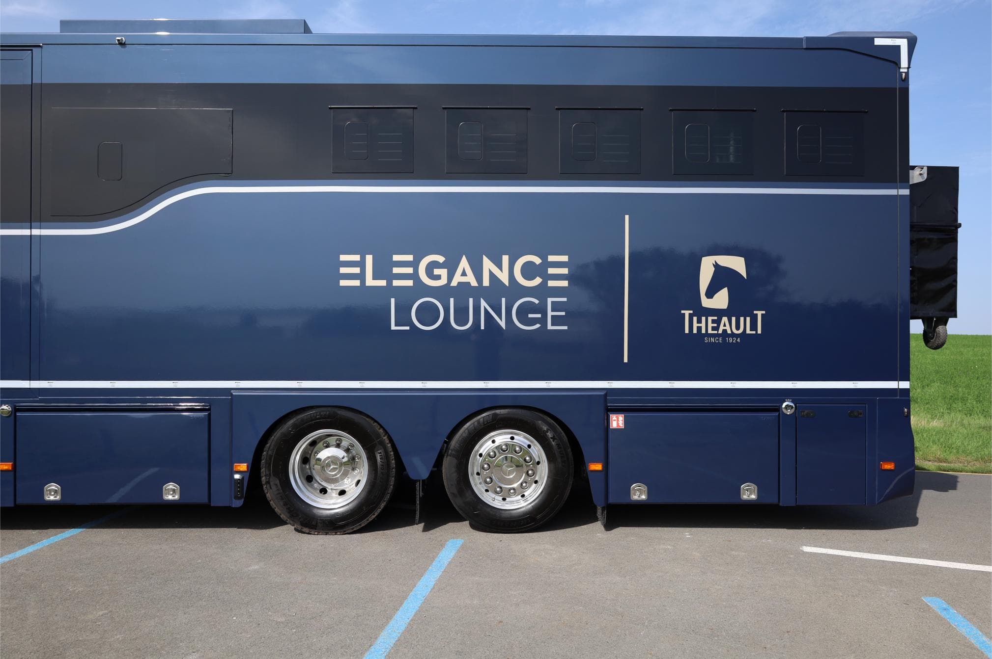 Vehículo azul "Elegance Lounge" con el logotipo de Theault, estacionado en la carretera asfaltada cerca de un campo verde.