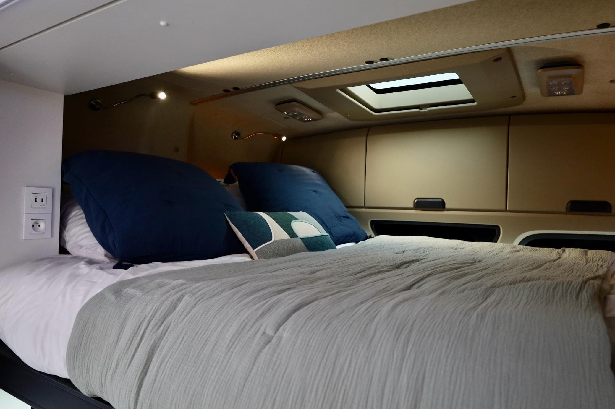 Acogedor interior de caravana con una cama cuidadosamente hecha, almohadas suaves, iluminación cenital y un tragaluz encima.