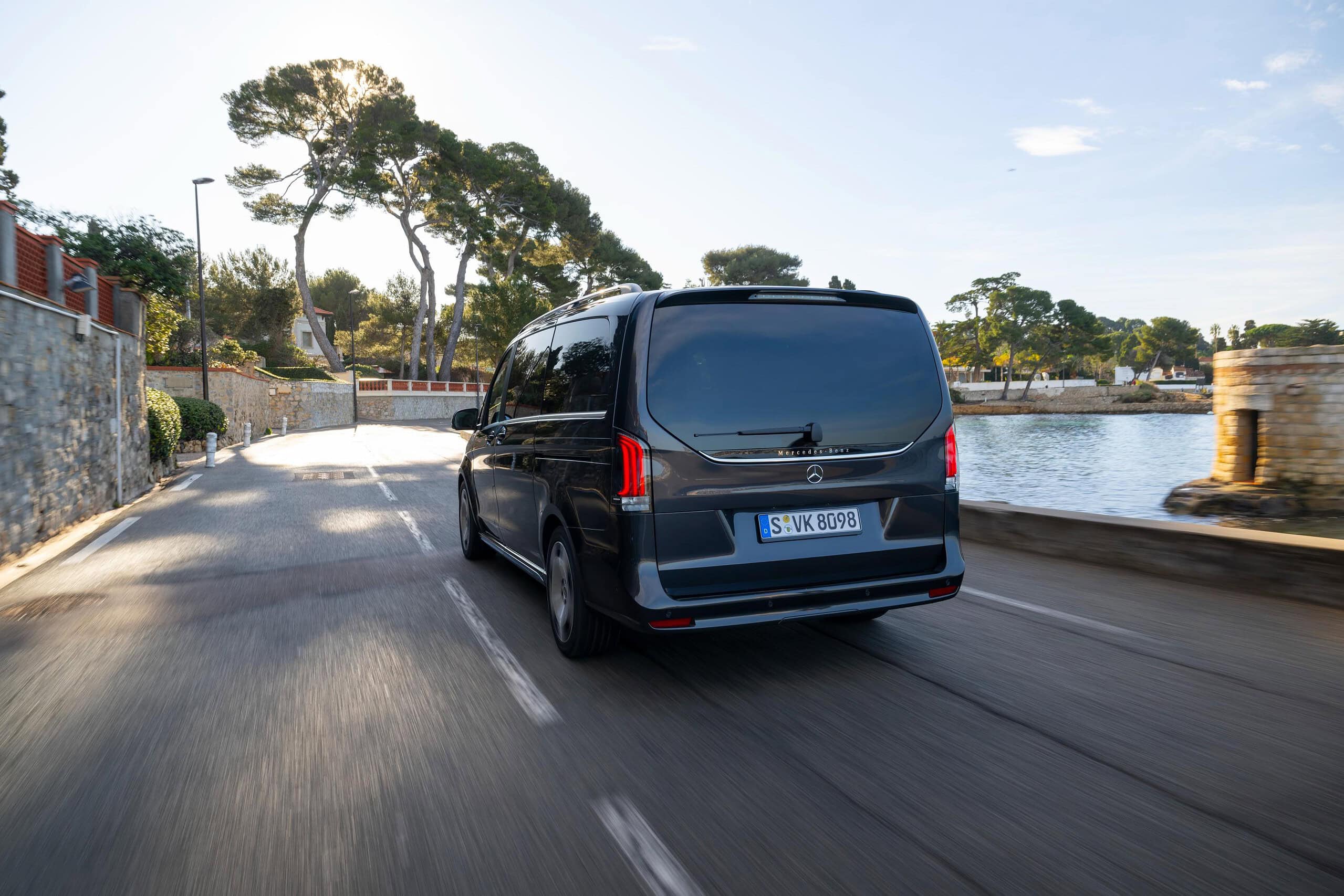 Una furgoneta Renault Master negra circula por una soleada carretera costera, una ecuación perfecta con árboles y un muro de piedra al fondo.