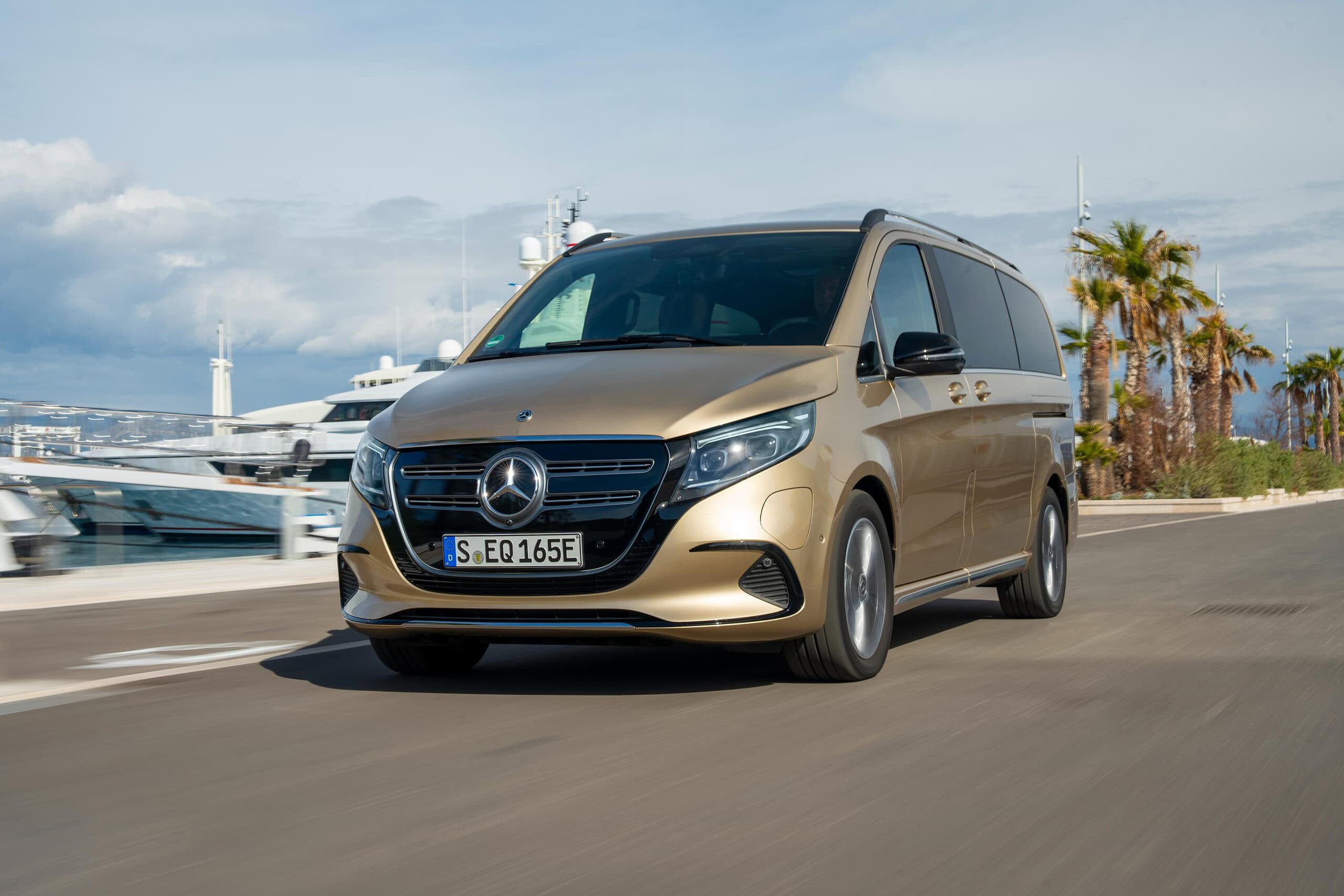 Una furgoneta Mercedes-Benz dorada circula por una carretera costera bordeada de palmeras y yates, creando la ecuación perfecta de lujo y ocio.