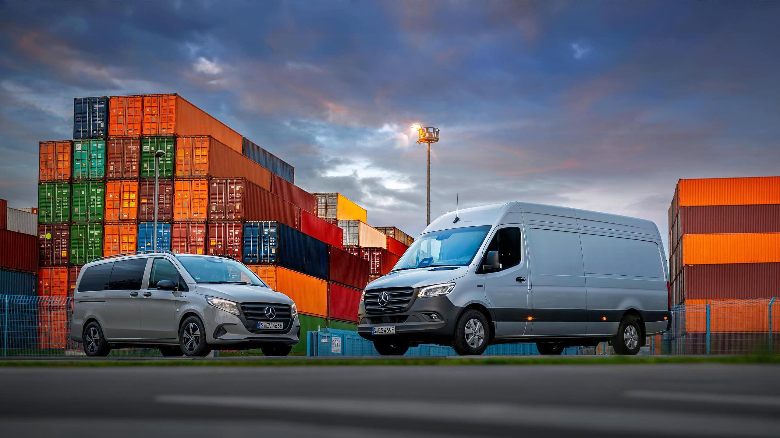 Al anochecer, dos furgonetas Mercedes-Benz están estacionadas frente a contenedores de envío coloridos apilados, creando la ecuación perfecta de tonos vibrantes y diseño elegante.