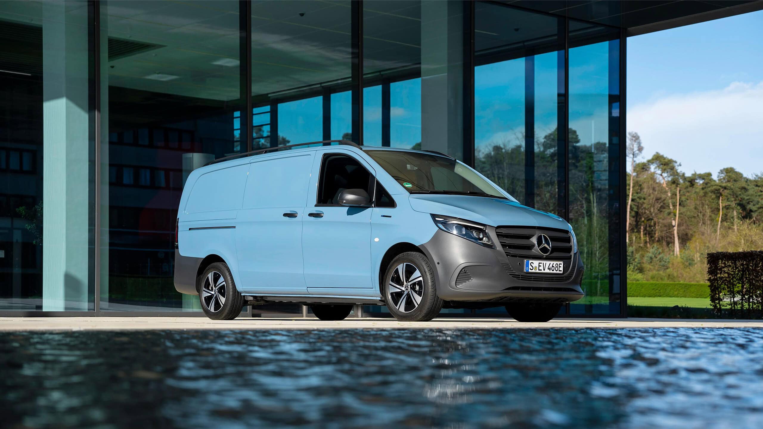 Una camioneta Mercedes-Benz azul claro está estacionada afuera de un moderno edificio de cristal cerca de agua reflectante, creando la ecuación perfecta de estilo y elegancia.