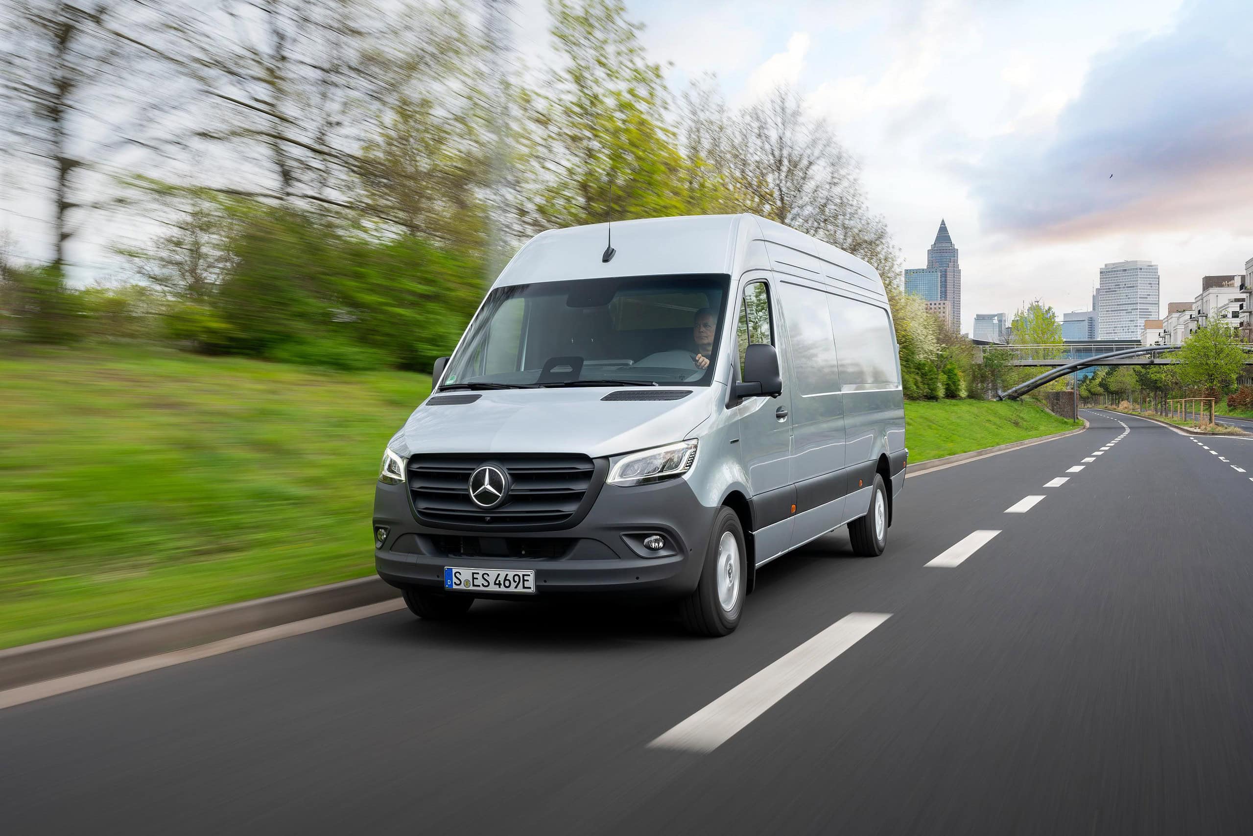 Una elegante furgoneta Mercedes-Benz plateada se desliza por la carretera, flanqueada por altos árboles y un horizonte urbano que forma la ecuación perfecta de la elegancia urbana.