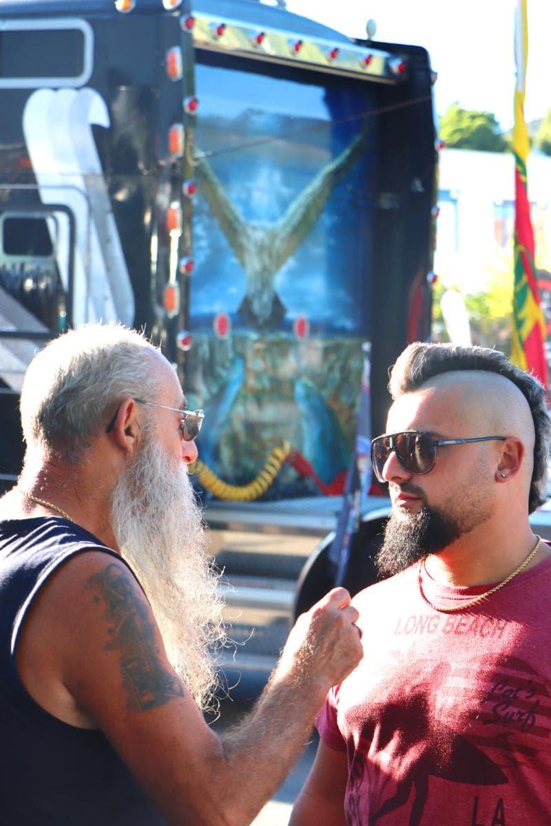 Dos hombres, uno con barba larga y el otro con gafas de sol, mantienen una conversación delante de un camión decorativo que parece ser un antiguo modelo de borrador automático.
