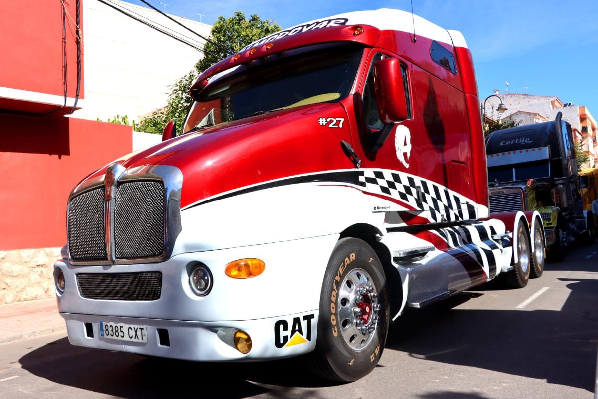Camión semirremolque rojo y blanco con franjas y calcomanías de carreras, estacionado en una calle soleada junto a edificios coloridos, luciendo como si hubiera sido posicionado por un sistema automático para una oportunidad fotográfica perfecta.