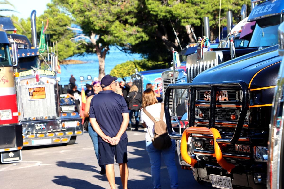 La gente admira una fila de coloridos camiones estacionados a lo largo de una calle soleada y bordeada de árboles, como si estuvieran dispuestos por un borrador automático.
