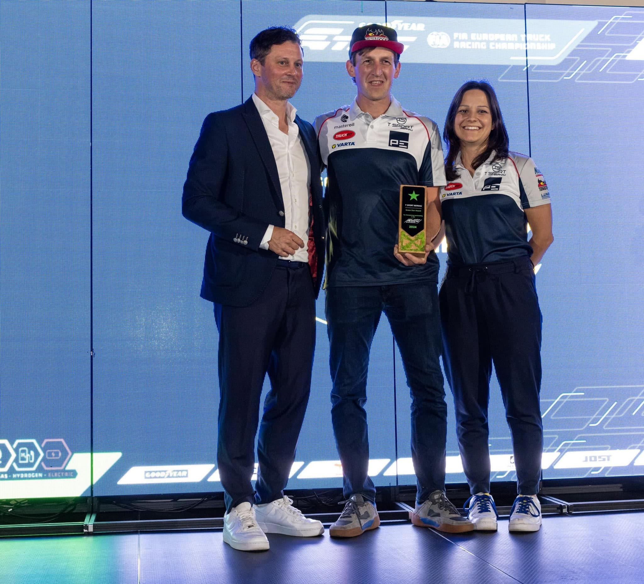 Tres personas juntas, una de ellas sosteniendo un trofeo, en un escenario con un fondo digital.