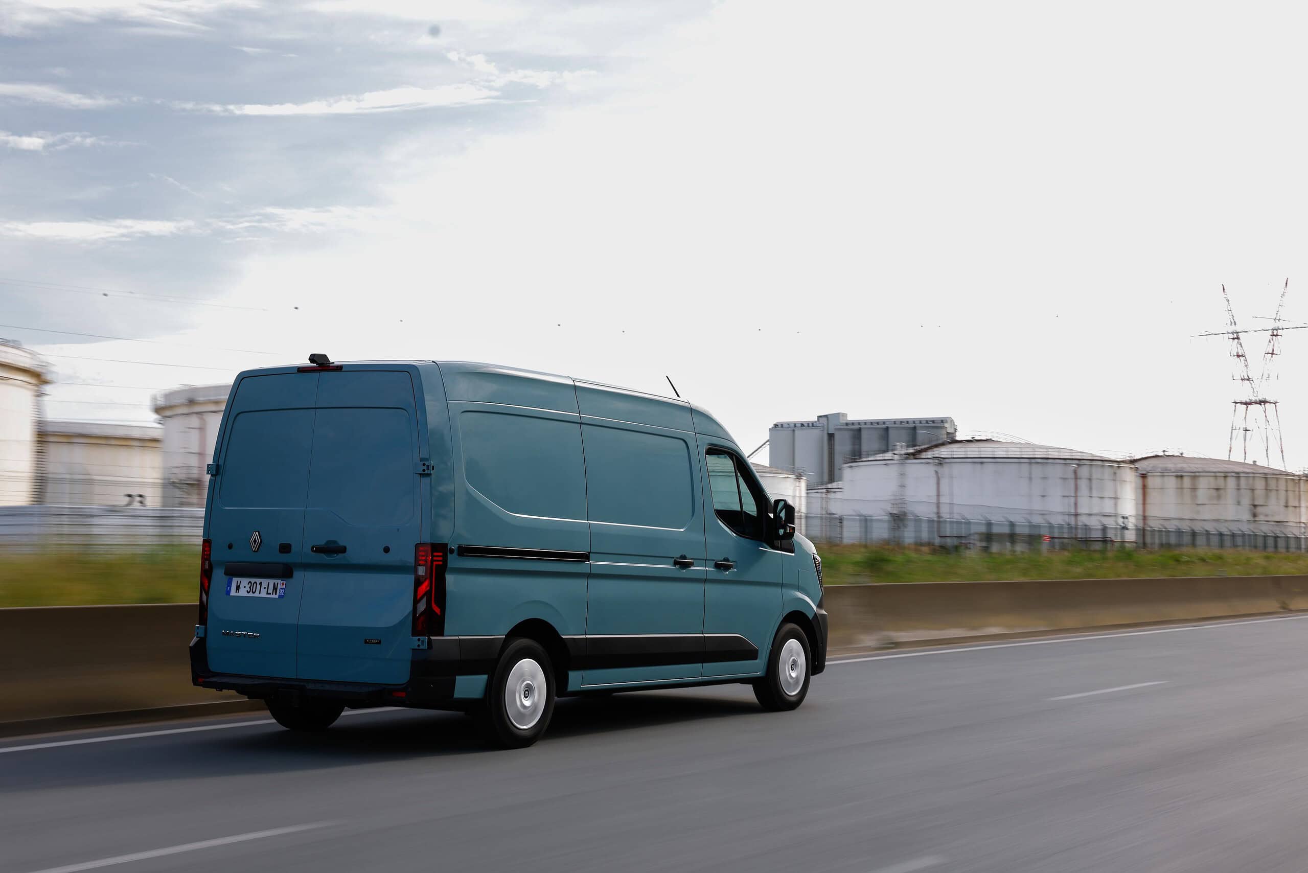 Una furgoneta Renault Master azul circula por la autopista pasando por edificios industriales, encarnando la ecuación perfecta bajo un cielo nublado.