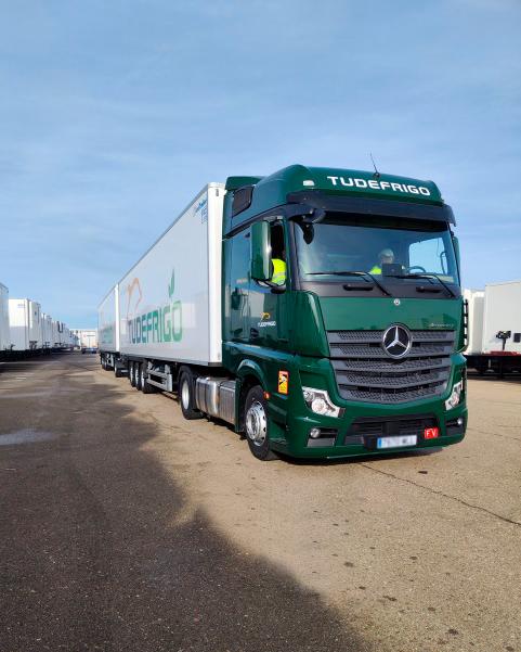 Un camión Mercedes TudeFrigo verde estacionado en una carretera industrial bajo un cielo despejado se mantiene firme para garantizar la seguridad en el transporte de mercancías, recordando a un potente duotráiler.