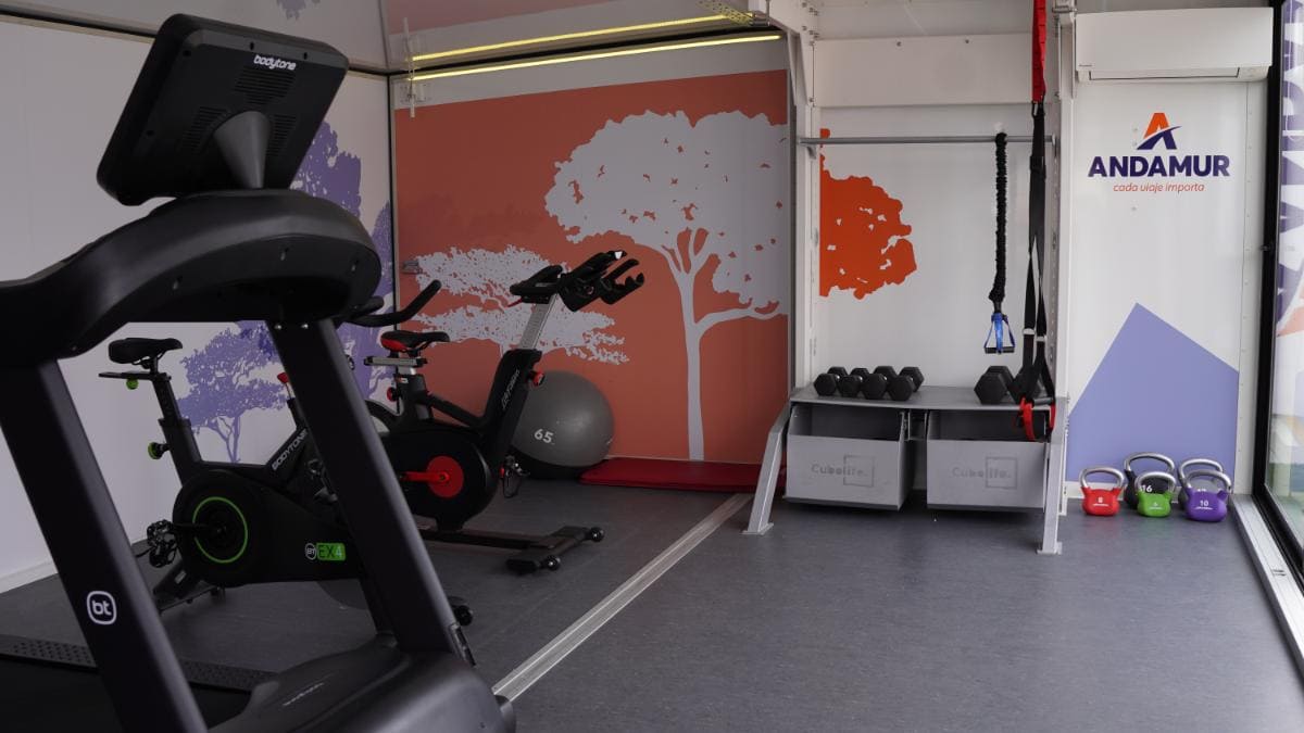 El interior de un gimnasio exhibe equipos de ejercicio, pesas rusas y vibrantes obras de arte en la pared con árboles, lo que crea un ambiente inspirador para su viaje de acondicionamiento físico. Embárquese en su ruta de salud con Andamur para energizar tanto el cuerpo como el espíritu en este espacio animado.