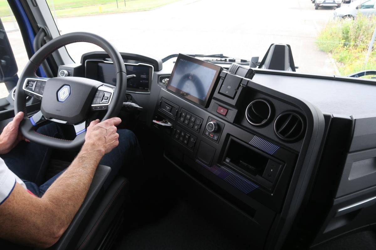 Persona conduciendo un camión Renault visto de lado, mostrando volante y tablero de instrumentos con múltiples paneles de control y una pantalla, indicativo de su idoneidad para distribución regional eléctrica.