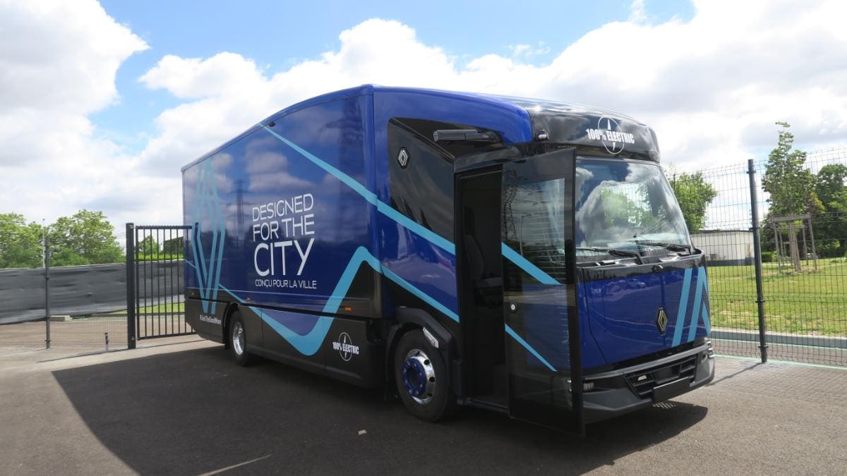 Un camión eléctrico azul, marcado con la palabra "Diseñado para la ciudad" en un lateral, se encuentra cerca de una valla. Este vehículo de Renault Trucks es ideal para la distribución regional eléctrica, ya que combina estilo y eficiencia para los paisajes urbanos.
