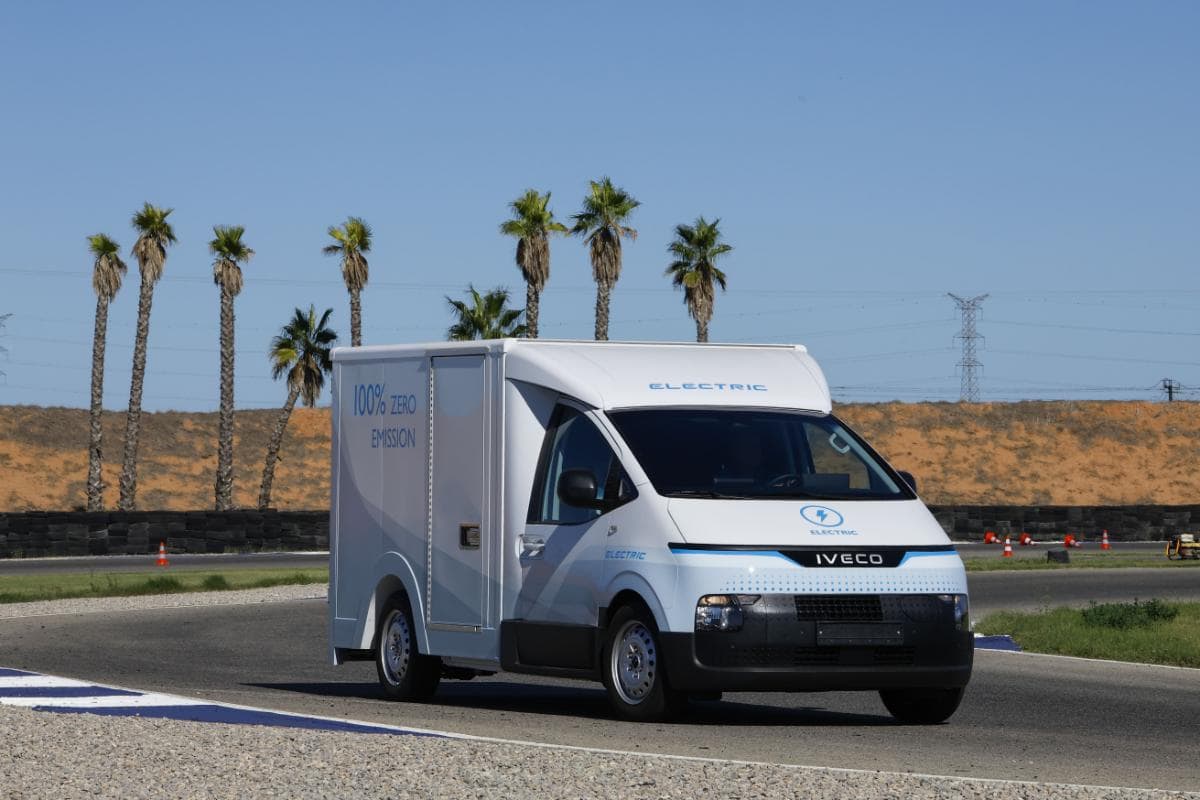 Una furgoneta eléctrica de reparto IVECO circula por una carretera soleada, enmarcada por palmeras ondulantes, encarnando la nueva gama de vehículos ecológicos perfectos para un transporte eficiente, imaginándola recorriendo rutas tan eficientemente como en el Circuito Aspar de Valencia.