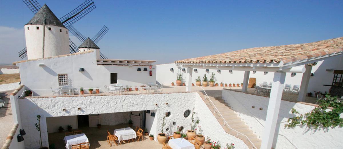 En un encantador patio que recuerda a La Mancha, las paredes blancas enmarcan mesas y plantas en macetas, mientras elegantes torres de molinos de viento se elevan hacia el cielo azul claro.