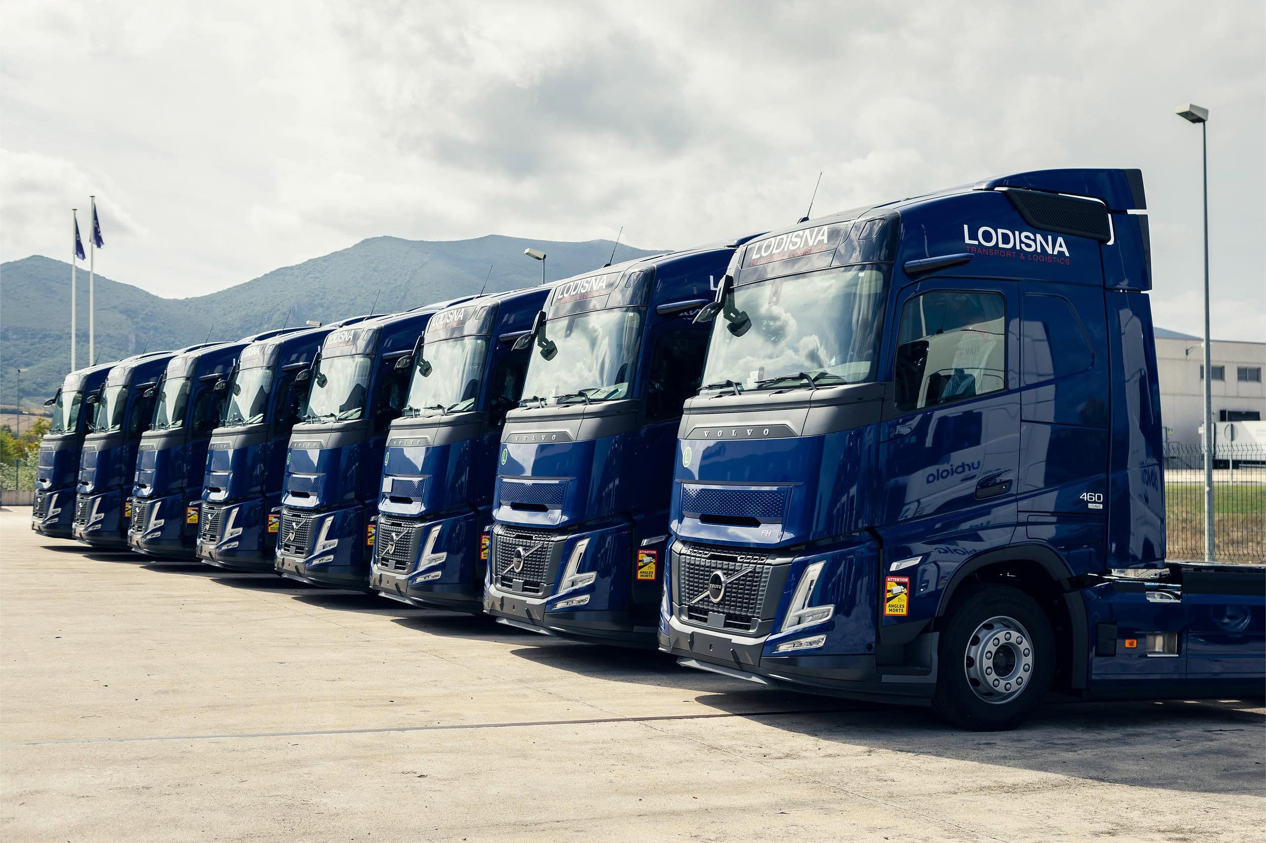 Una fila de camiones Volvo azules, que cuentan con la tecnología I-Save de bajo consumo de combustible, están estacionados en un lote abierto con majestuosas montañas de fondo.