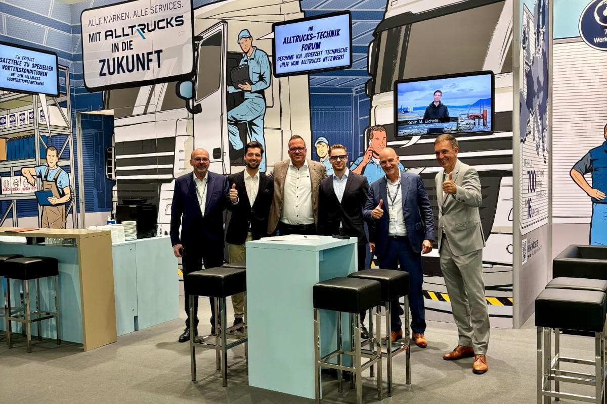 Un grupo de seis hombres vestidos de negocios posan en un stand de una feria comercial, con un fondo de camiones como tema. La escena rezuma profesionalidad e innovación, como si estuvieran preestrenando lo último en tecnología de elaboración automática de cartas.