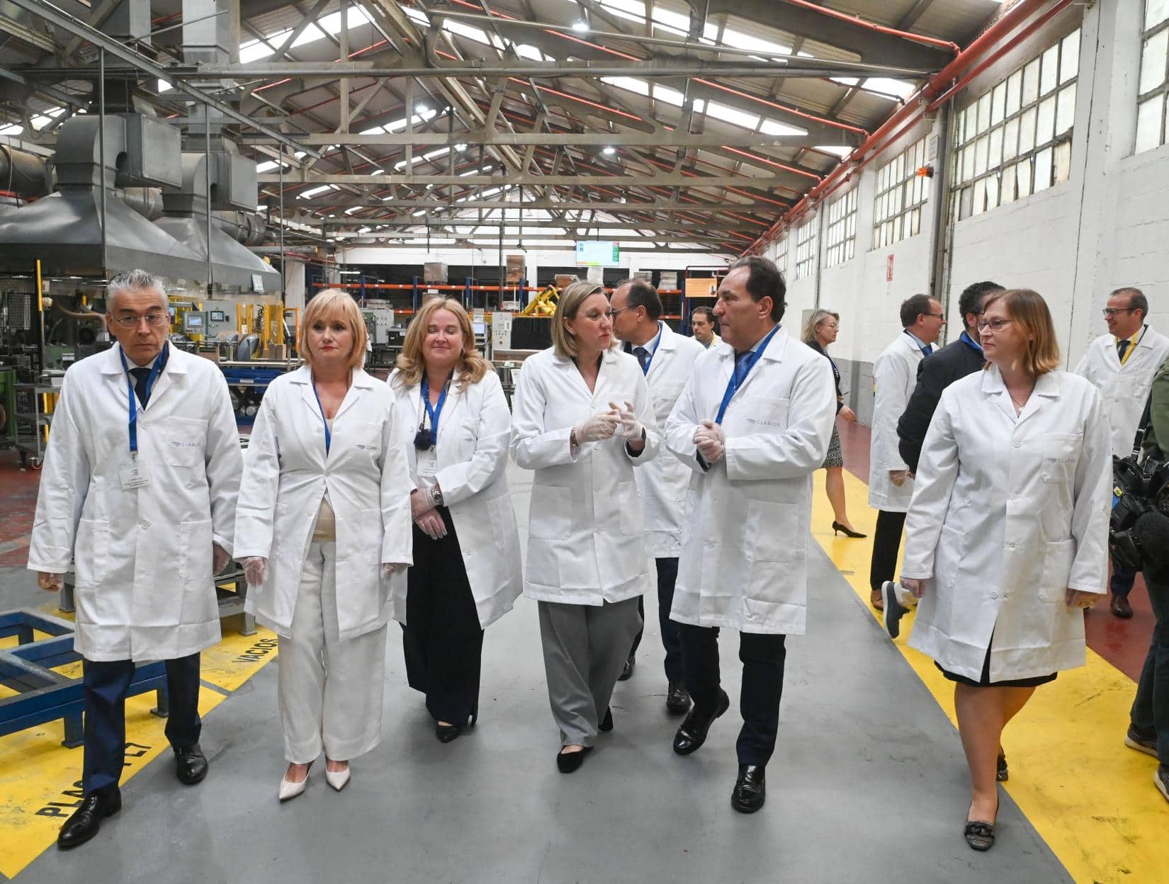 Personas con batas de laboratorio y placas de identificación caminando por una instalación industrial con maquinaria al fondo.