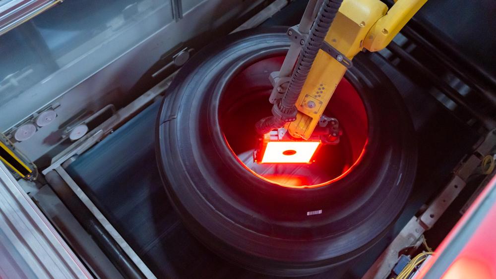 Bajo el resplandor de la iluminación roja en un entorno industrial, un brazo robótico utiliza su función de borrador automático para inspeccionar meticulosamente un neumático, garantizando precisión y eficiencia.