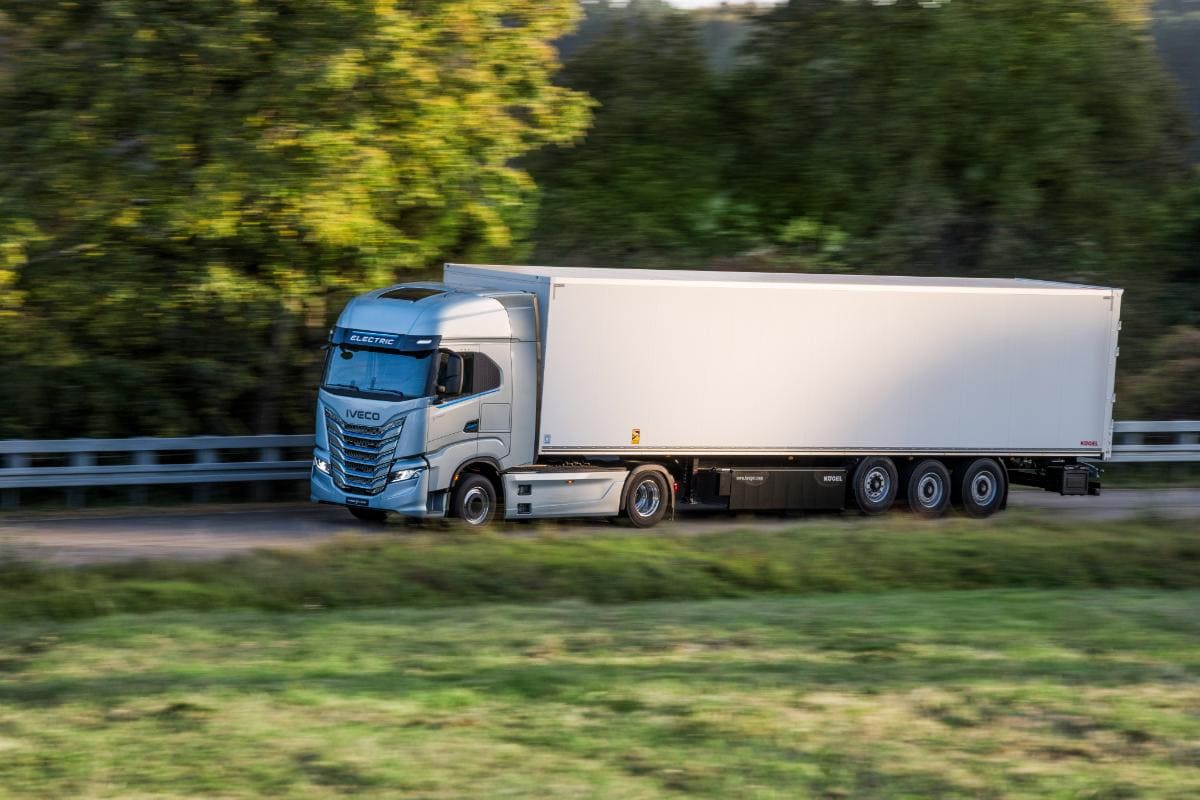 Un camión eléctrico azul y blanco, testimonio de la revolución de la electromovilidad, circula por una carretera con una exuberante vegetación de fondo, encarnando el espíritu de innovación que prospera en toda España mientras se enfrenta al desafío del transporte sostenible.