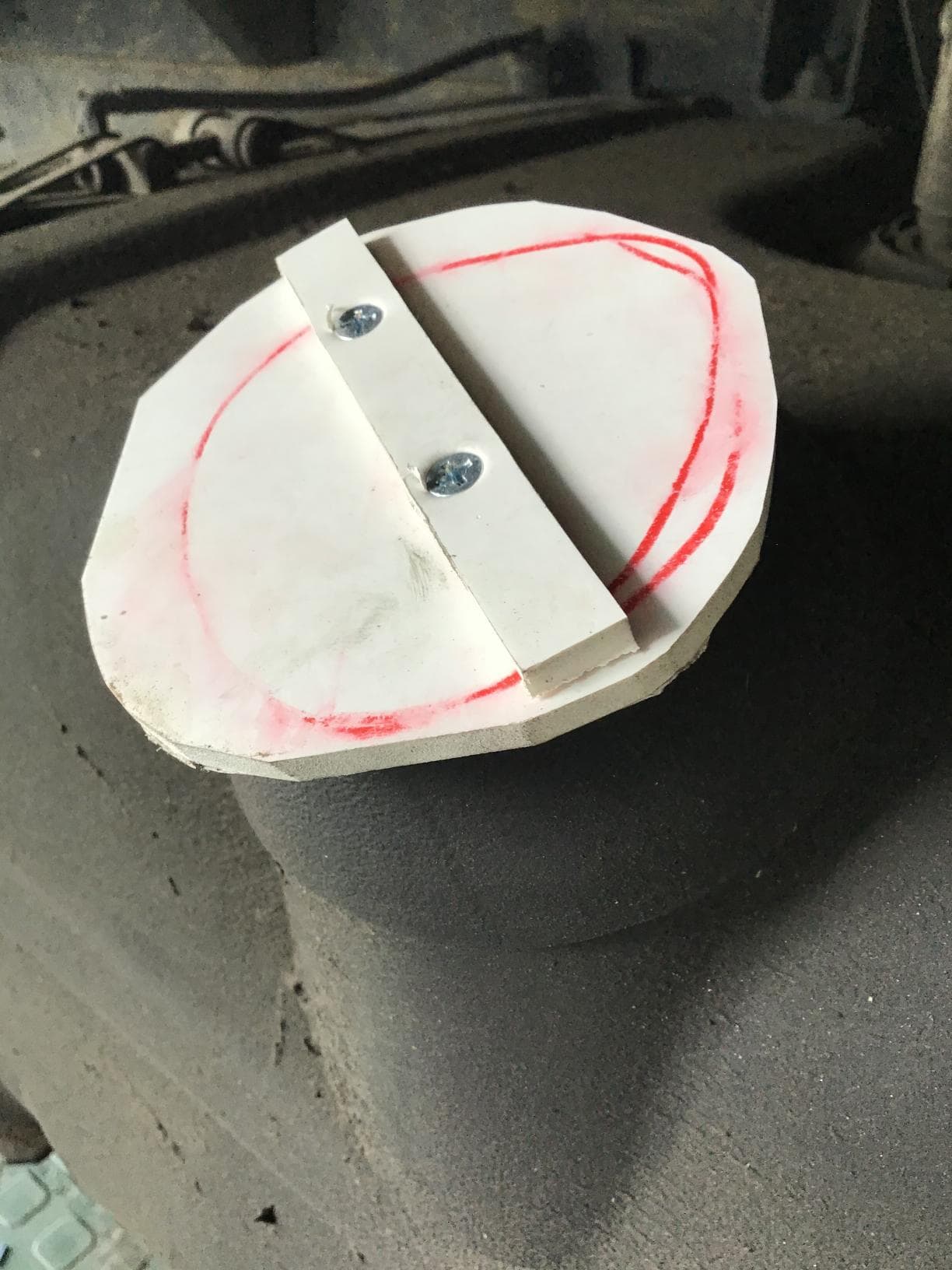 Una tapa de plástico blanca con dos tornillos sobre un objeto cilíndrico grande, marcado con un anillo rojo.