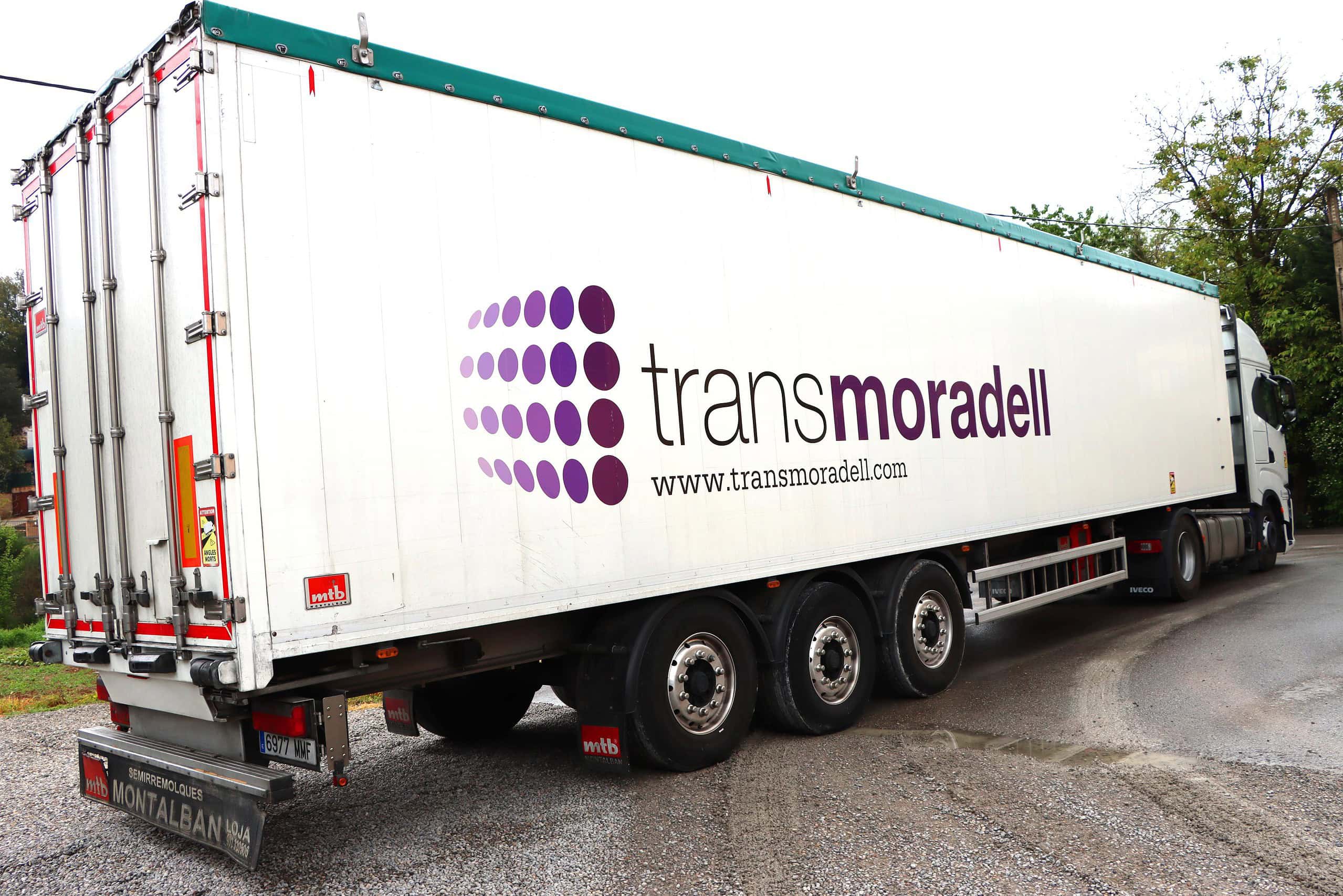 Un gran camión con el logo “transmoradell” está estacionado en una carretera cerca de árboles, simbolizando el compromiso de la empresa de crecer y transportar sueños a través de las distancias.