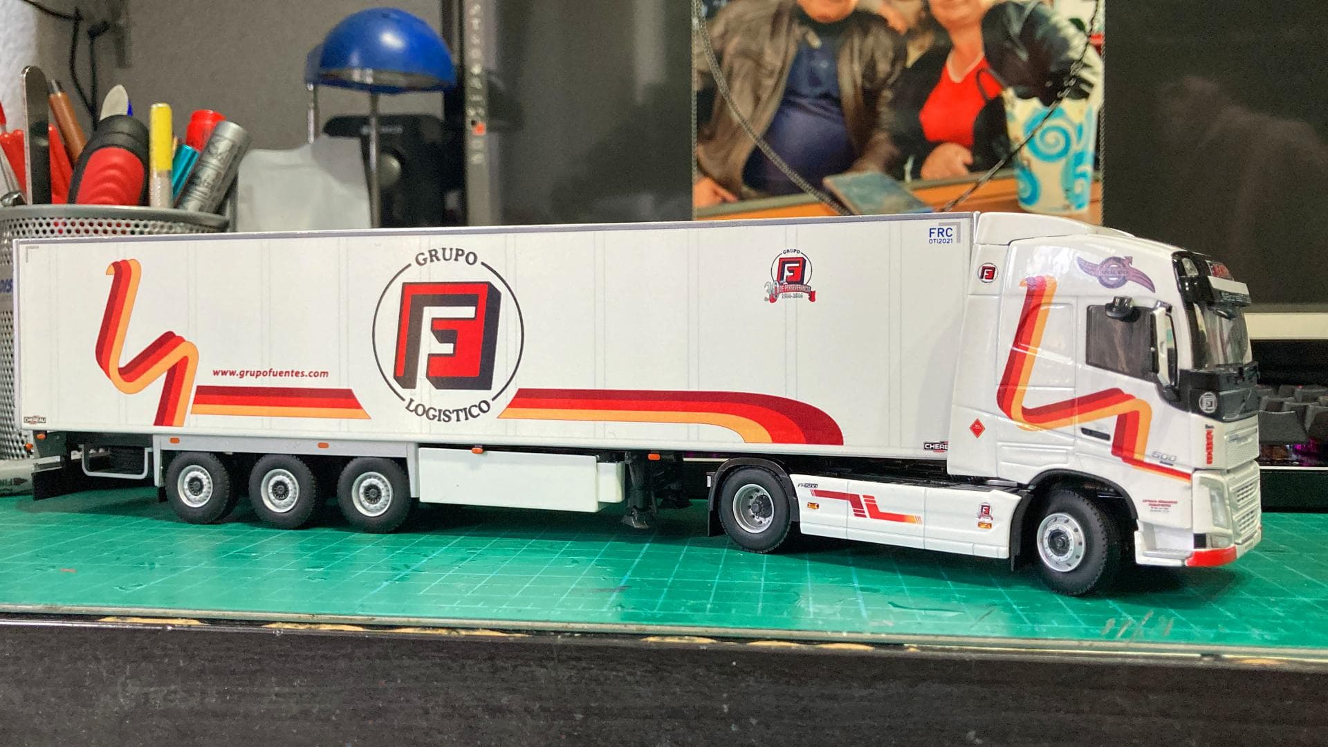 Maqueta de un camión semirremolque blanco con el logo de "Grupo Logístico", sobre una estera de corte verde con herramientas de trabajo al lado. Recuerda el recorrido histórico de Antonio Fernández por caminos desafiantes, superando innumerables obstáculos en la industria logística.