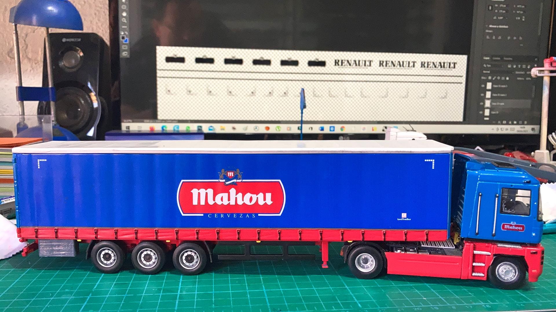 El camión modelo con el logo “Mahou” en su remolque azul se encuentra junto a un ordenador que muestra un software de diseño, reflejando el recorrido de Antonio Fernández a través de los obstáculos creativos en su camino hacia la artesanía.