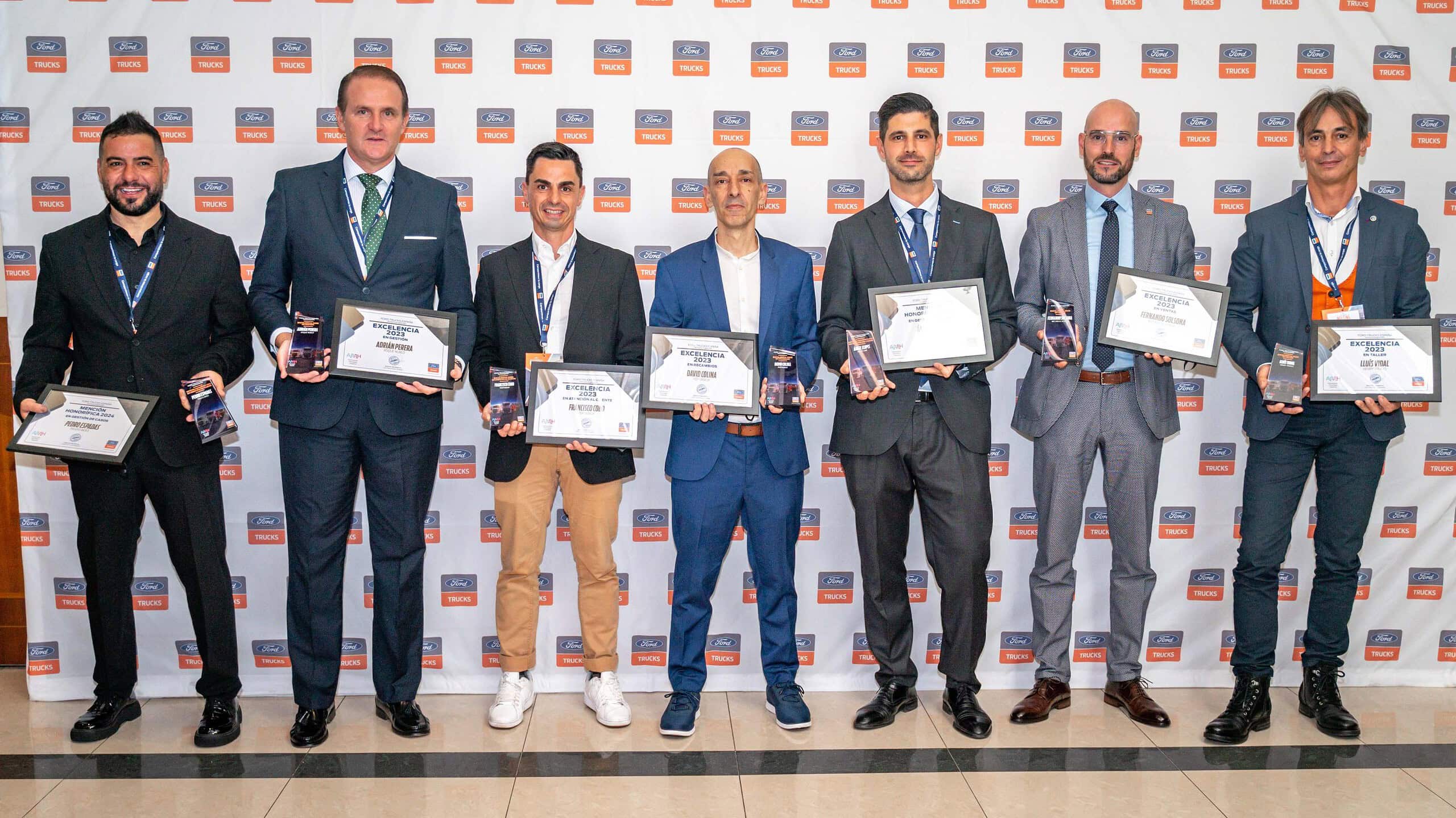 Siete hombres vestidos de traje y con premios en la mano se paran orgullosos frente a un fondo cubierto de logotipos, celebrando en la Gala de la Excelencia en España.