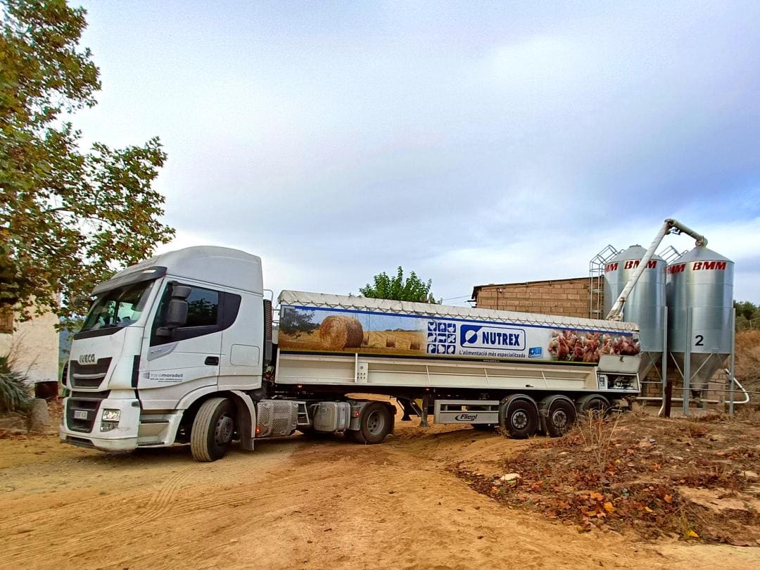 El camión blanco Nutrex de TransMoradell estaba estacionado cerca de silos de almacenamiento en un camino de tierra bajo un cielo nublado.