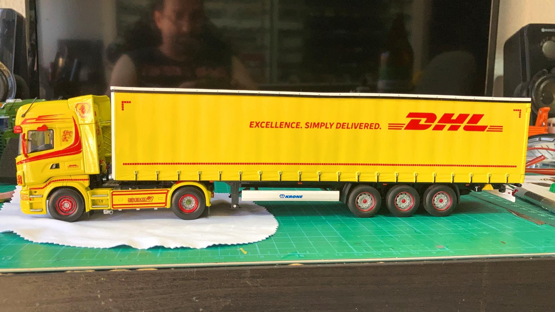 Camión modelo DHL con remolque amarillo y texto en rojo: “EXCELENCIA. SIMPLEMENTE ENTREGADA.” sobre una alfombra verde, en posición vertical como el camino de Antonio Fernández, superando obstáculos a cada kilómetro.