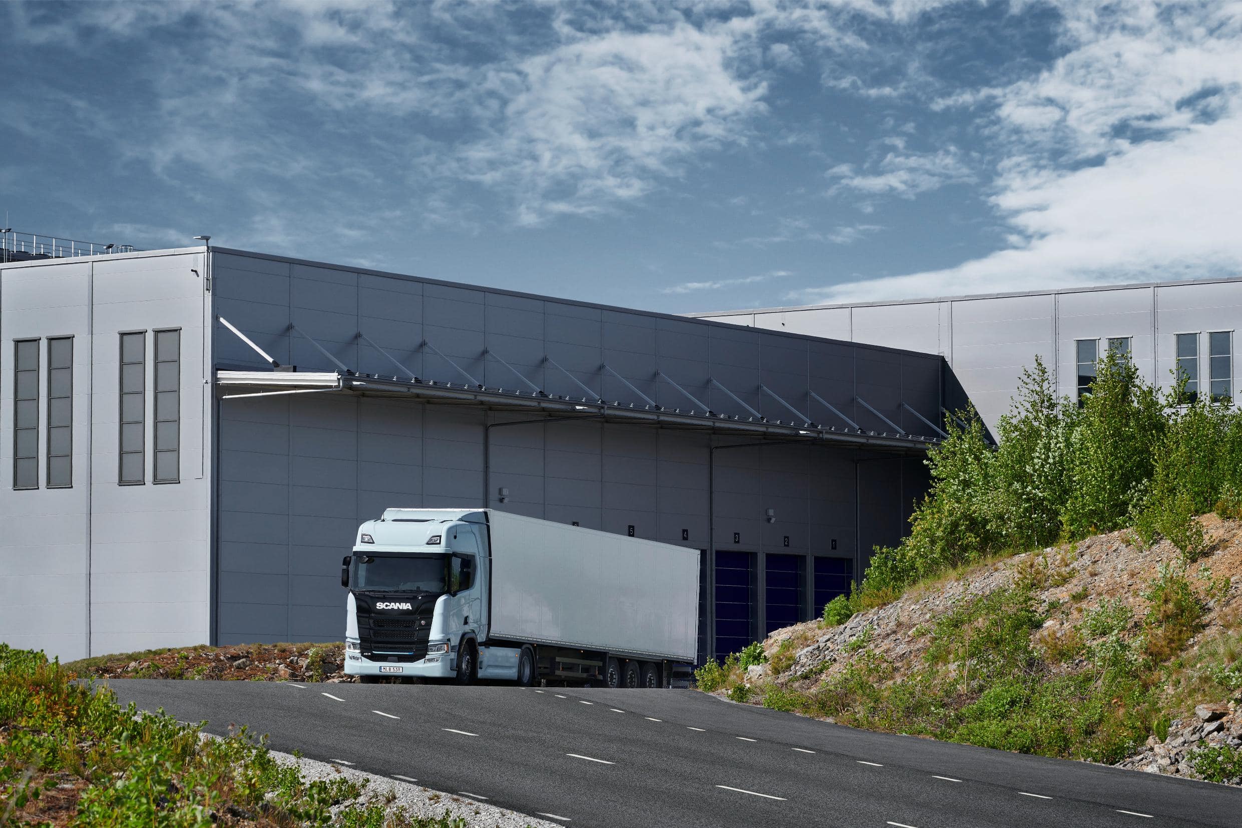 Un camión eléctrico blanco está estacionado en un gran edificio industrial en un día claro, con una exuberante vegetación y una carretera en primer plano, mostrando el futuro del transporte sostenible.