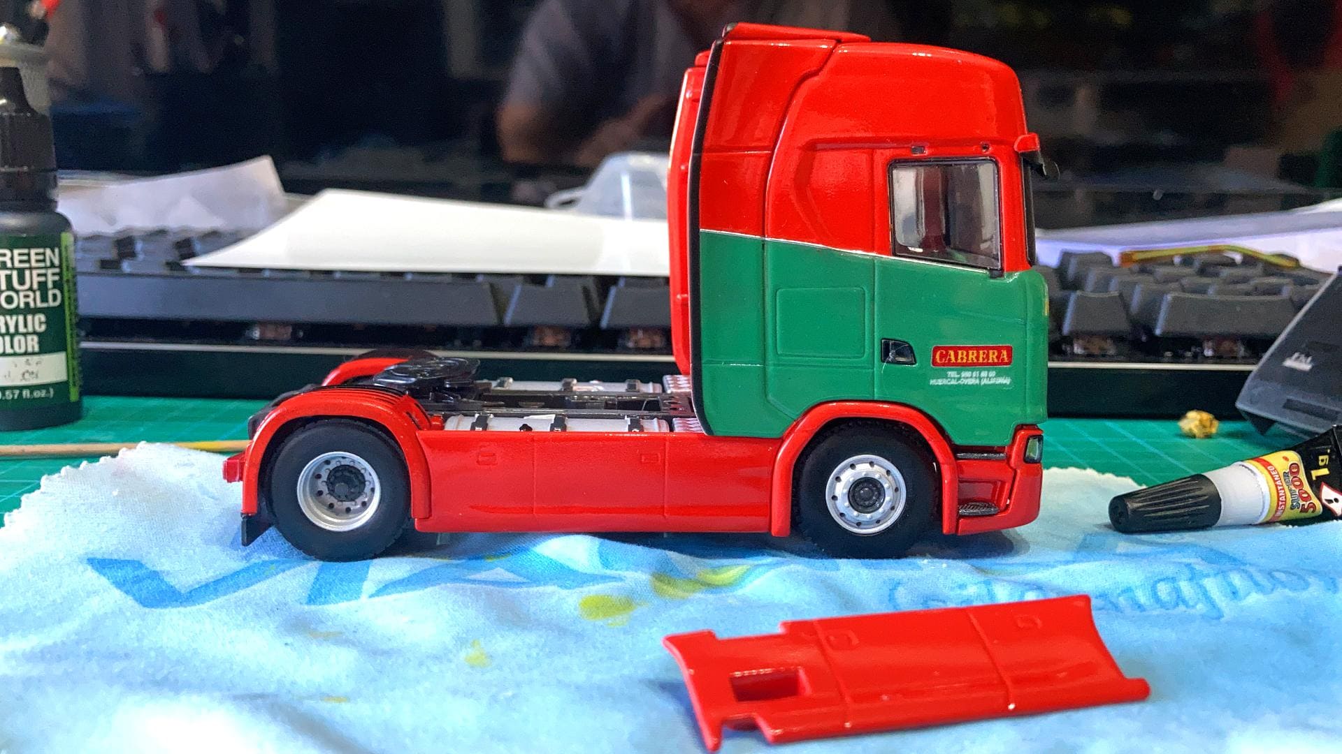 Sobre un banco de trabajo se construye una maqueta de un camión rojo y verde que recuerda a los diseños de Antonio Fernández. Herramientas y materiales llenan el fondo, simbolizando el camino para superar los obstáculos creativos.
