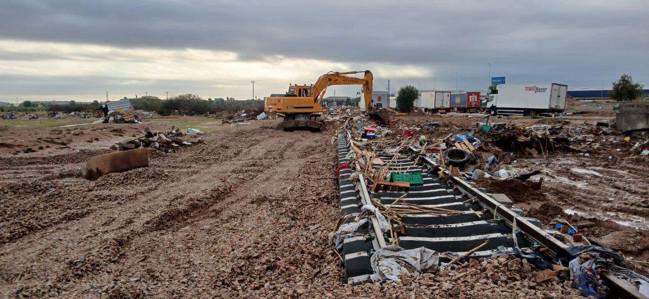 En una obra en construcción cerca de Valencia se ve una excavadora limpiando escombros y piedras bajo un cielo nublado, preparando el área para nuevas vías ferroviarias.