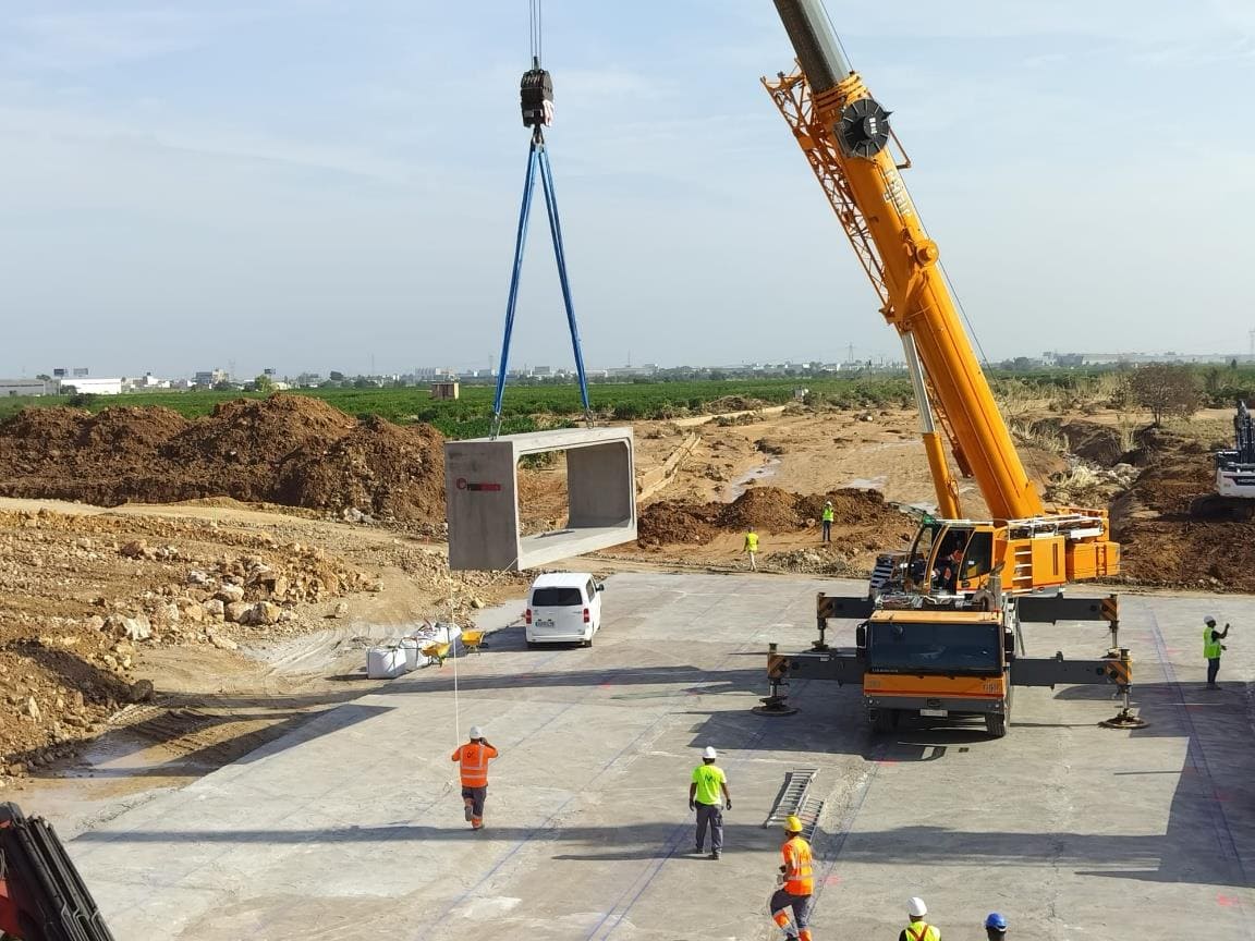 En una obra en plena actividad en Valencia, una grúa levanta con cuidado una gran viga de hormigón, con trabajadores y vehículos cerca. La precisión de la operación garantiza la integración perfecta de esta pieza vital en la infraestructura en desarrollo que rodea las vías ferroviarias de la ciudad.