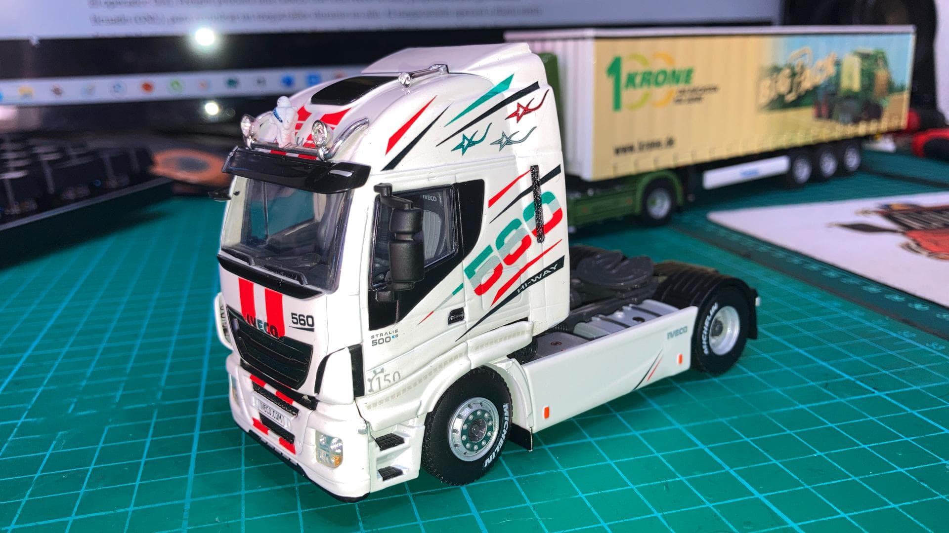 Camión modelo con diseño en blanco y rojo sobre tapete de corte verde, que recuerda al estilo de Antonio Fernández, navegando su camino junto al remolque del fondo.
