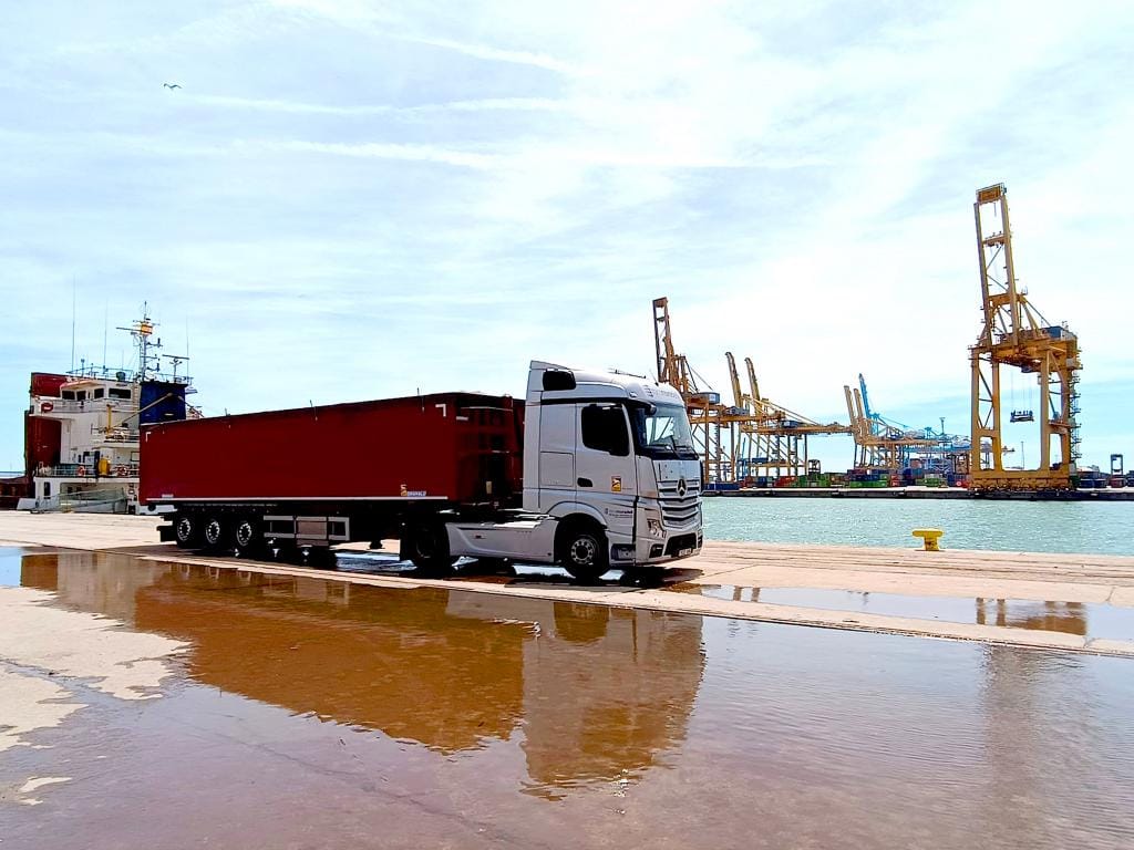 Un camión de TransMoradell está estacionado cerca de un bullicioso puerto de envío, con grúas y contenedores de carga coloridos en el fondo en un día soleado, listo para crecer y crear oportunidades en el comercio global.