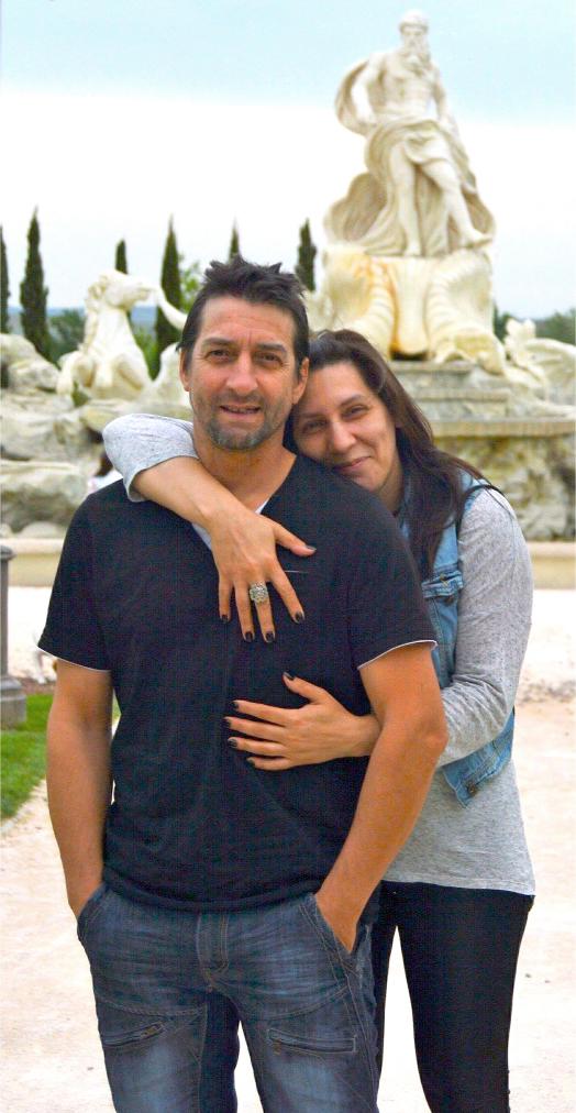 Una pareja abrazándose cálidamente frente a una majestuosa fuente al aire libre con estatuas, que recuerdan el arte de Antonio Fernández, simboliza su viaje superando obstáculos en el camino de la vida.