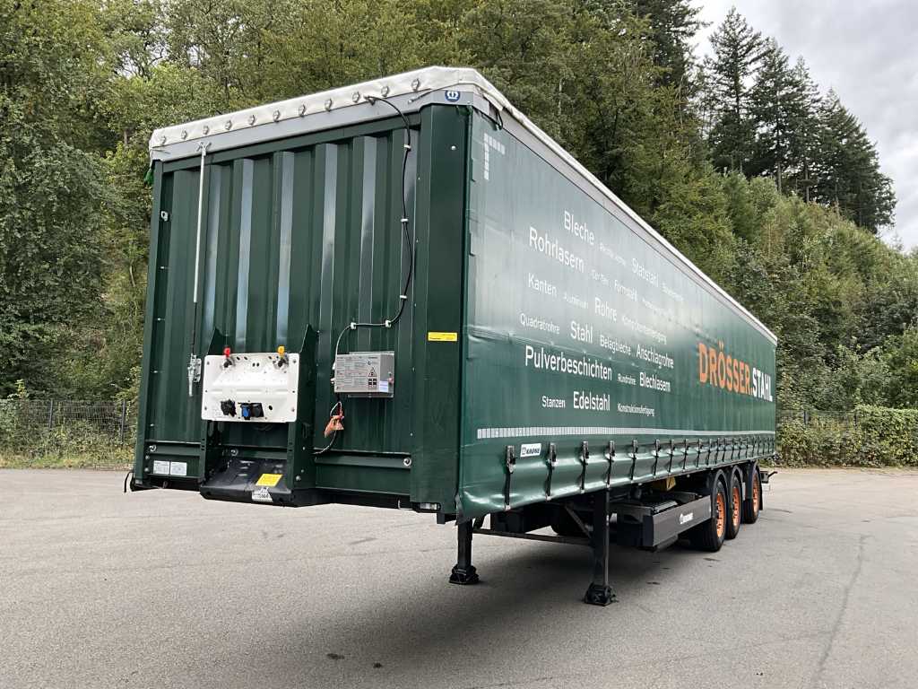 Un remolque de camión verde, que a menudo se ve transportando camiones, está estacionado en una carretera cerca de árboles, con un texto blanco en negrita en su costado.
