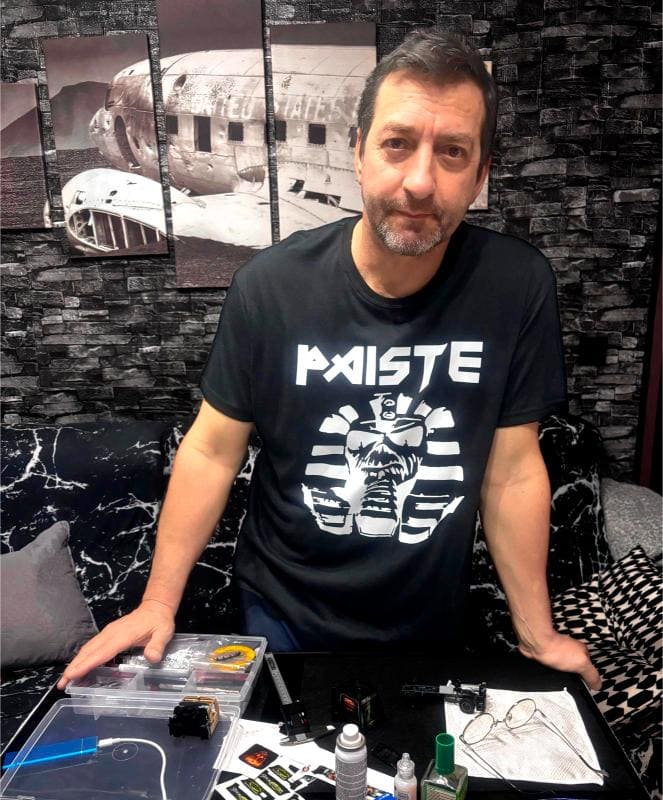 Antonio Fernández, con una camiseta negra de "PAISTE", se encuentra junto a una mesa con material para tatuar en una sala marcada por su decoración en blanco y negro. Su pasión es un testimonio de sabiduría en construcción.