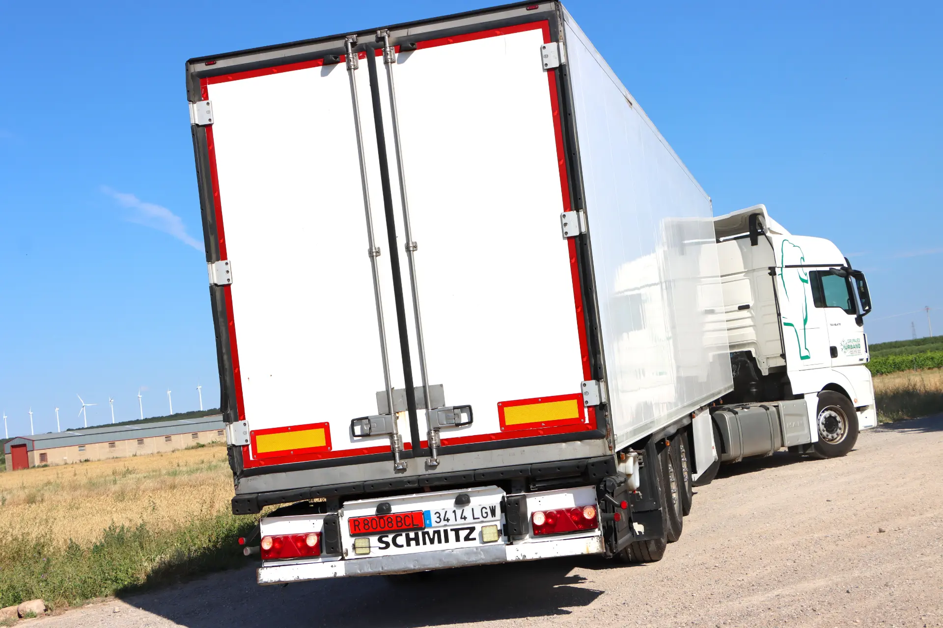 Una camioneta blanca y un remolque recorren un camino rural de tierra bajo el vasto cielo azul, con turbinas eólicas girando suavemente en el fondo.